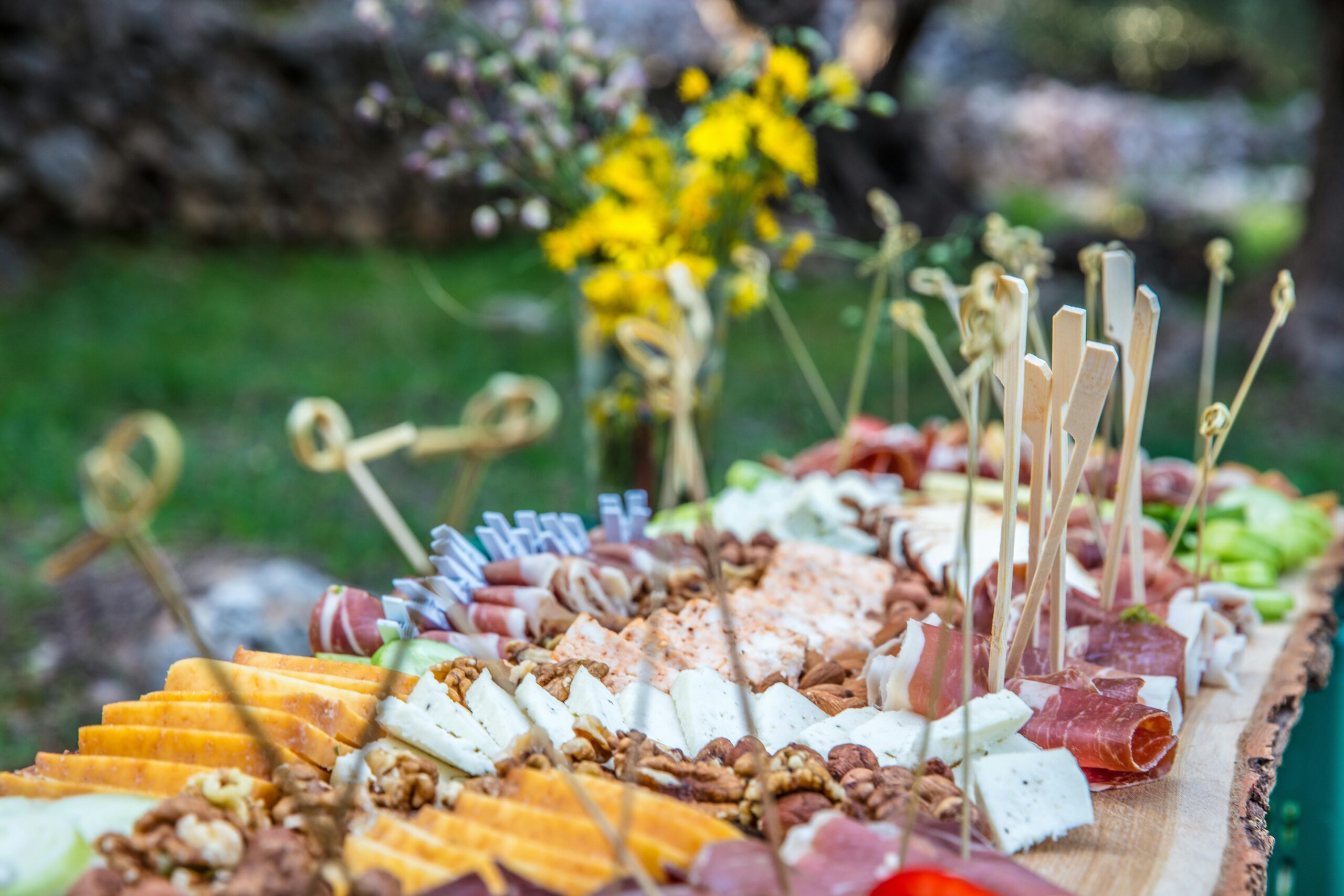 Plateau de charcuterie et fromage avec brochettes, fleurs floues en arrière-plan.